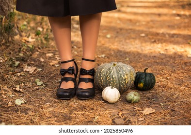 Happy Halloween! Cropped Photo Of Female Feet In Retro Leather Black Shoes With Bows, Standing On Drowning Path Of Dry Leaves And Grass Next To Multi-colored Pumpkins Outside In Autumn Sunny Day