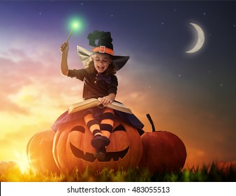 Happy Halloween! Cute Cheerful Little Witch With A Magic Wand And Book Of Spells. Beautiful Child Girl In Witch Costume Sitting On The Big Pumpkin, Conjuring And Laughing.