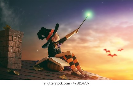 Happy Halloween! Cute Cheerful Little Witch With A Magic Wand And Book Of Spells. Beautiful Child Girl In Witch Costume Sitting On The Roof, Conjuring And Laughing.