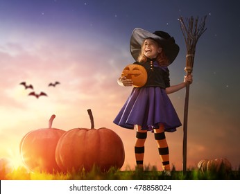 Happy Halloween! Cute Little Witch With A Big Pumpkin. Beautiful Young Child Girl In Witch Costume Outdoors. 