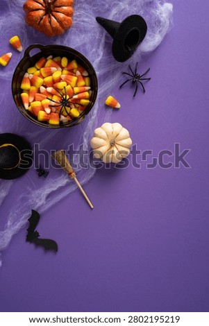 Happy Halloween flatlay featuring candy corn in a bowl, pumpkins, witch hat, broom, spiders and cobweb on a purple background