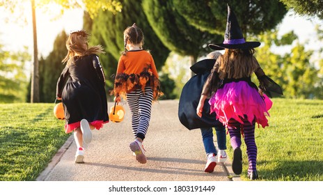 Happy Halloween! Funny Kids Girls Laughing In Fancy Dress With Bucket Run Back Along The Path In The Garden Outdoor
