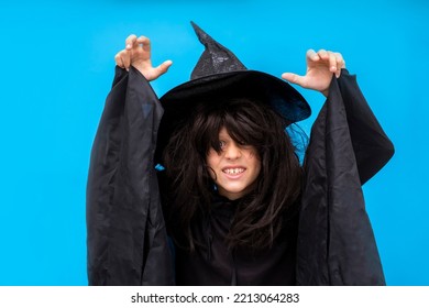 Happy Halloween Kid Raises His Hands Up While Casting A Spell. Child In Black Sorcerer's Costume. Witch On Blue Background. Boy In A Suit Wizard. Copy Space Trick Or Treating