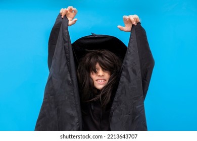 Happy Halloween Kid Raises His Hands Up While Casting A Spell. Child In Black Sorcerer's Costume. Witch On Blue Background. Boy In A Suit Wizard. Copy Space Trick Or Treating