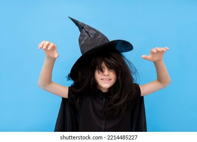 Happy Halloween Kid Raises His Hands Up While Casting A Spell. Child In Black Sorcerer's Costume. Witch On Blue Background. Boy In A Suit Wizard. Copy Space Trick Or Treating