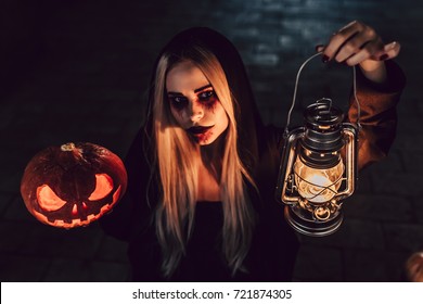 Happy Halloween! Portrait of black witch with scary makeup in black mantle. Holding pumpkin and lamp in hands - Shutterstock ID 721874305