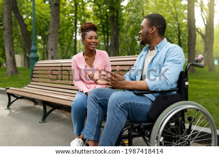 Happy handicapped black guy in wheelchair talking to his girlfriend at park, copy space. Young African American woman sitting on bench, communicating to her disabled male friend outdoors