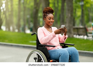 Happy handicapped black woman in wheelchair using smartphone, checking messages, browsing internet at city park. African American disabled lady with mobile device posting in social network outdoors