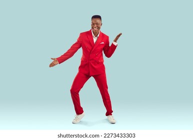 Happy handsome African American guy in stylish red suit dancing in fashion studio. Joyful attractive Tanzanian man in funky jacket and trousers dancing to lively music on light blue colour background