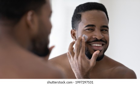 Happy handsome African American man applying moisturizer on face, putting collagen anti age cream, sunscreen, cleansing lotion, caring for hydrating soft skin, reducing wrinkles, acne, eyebags - Powered by Shutterstock