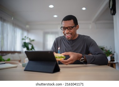 Happy Handsome African American Man Is Holding A Hamburger Celebrating Birthday Through Video Call Virtual Party With Friends By A Tablet Distance At Home. Modern Holiday Celebration