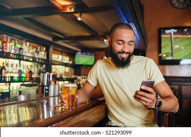 Happy handsome african american young man drinking beer in bar and using smartphone  - Powered by Shutterstock