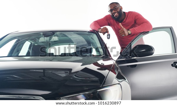 Happy handsome African man showing car keys in near\
his newly bought vehicle car smiling cheerfully copy space owner\
ownership sales driving consumerism private taxi concept studio\
shot