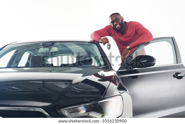 Happy handsome African man showing car keys in near\
his newly bought vehicle car smiling cheerfully copy space owner\
ownership sales driving consumerism private taxi concept studio\
shot