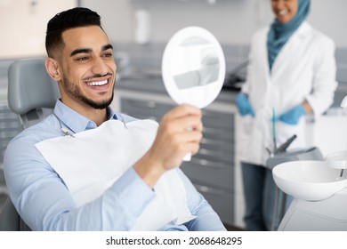 Happy Handsome Arab Man Looking To Mirror After Teeth Treatment In Modern Clinic, Middle Eastern Male Patient Sitting In Chair In Stomatological Cabinet And Enjoying His New Smile, Closeup - Powered by Shutterstock