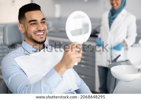 Happy Handsome Arab Man Looking To Mirror After Teeth Treatment In Modern Clinic, Middle Eastern Male Patient Sitting In Chair In Stomatological Cabinet And Enjoying His New Smile, Closeup