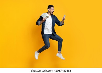 Happy Handsome Arabic Guy With Cash And Mobile Phone Jumping Up On Yellow Studio Background, Middle-eastern Young Man Gambling Or Trading Online, Using Mobile App, Full Length Shot