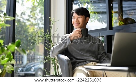 Happy and handsome Asian male enjoys having morning coffee at the coffee shop, looking away from camera, daydreaming about his career success.