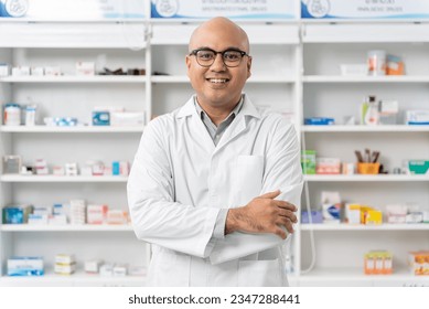 Happy handsome asian male pharmacist wearing eyeglasses and lab coat standing with arms crossed and looking at camera, He feels good, trustworthy and proud of his work in the pharmacy drugstore.