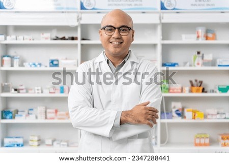 Happy handsome asian male pharmacist wearing eyeglasses and lab coat standing with arms crossed and looking at camera, He feels good, trustworthy and proud of his work in the pharmacy drugstore.