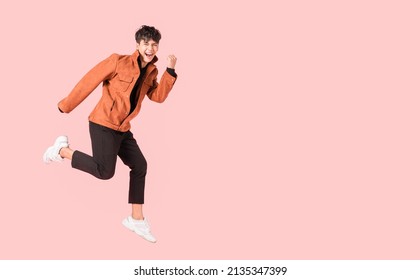 Happy Handsome Asian Man In Fashionable Clothing And Jumping Doing Winner Gesture Isolated On Pink Background. Portrait Of Young Male Cheerful Confident And Excited Jump On Air And Smile In Studio.