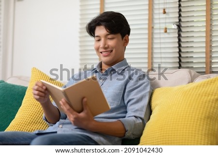 Happy handsome asian man sit on couch smile and reading book relax time at home.Cheerful businessman dressed in blue shirt sitting on sofa and reading a book