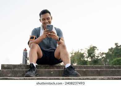 A happy and handsome Asian man in sportswear is sitting on the stairs and chatting with his friends on his phone after exercising running in a park. - Powered by Shutterstock