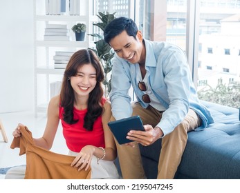 Happy Handsome Asian Man And Young Beautiful Woman Laughing Together While Looking At Tablet Screen. Woman Hold A Shirt And Man Holding Tablet On Couch Near The Glass Window At White Living Room.