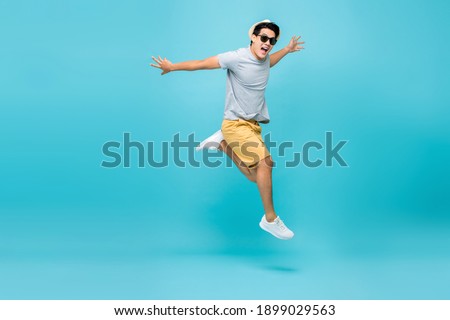 Happy handsome Asian tourist man in casual attire jumping studio shot isolated on light blue background