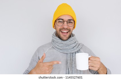 Happy handsome bearded male with white cup of tea wearing yellow hat and gray sweater with scarf in autumn or winter. Cheerful smiling man advertising something on white mug. Healthy, medicine, people - Powered by Shutterstock