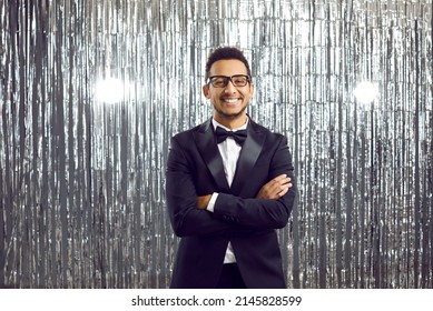 Happy Handsome Black Man In A Modern Tuxedo, White Shirt, Bowtie And Glasses Attending A Party. Smiling African Gentleman In An Elegant Suit Standing With His Arms Crossed On A Shiny Studio Background