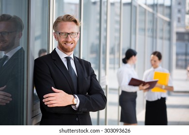 Happy Handsome Businessman With Colleagues Behind Him. Blond Man In Glasses And Black Business Suit Smiling With His Arms Crossed.