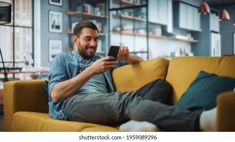 Happy Handsome Caucasian Man Using Smartphone in Cozy Living Room at Home. Man Resting on Comfortable Sofa. He's Browsing the Internet and Checking Videos on Social Networks and Having Fun. - Powered by Shutterstock