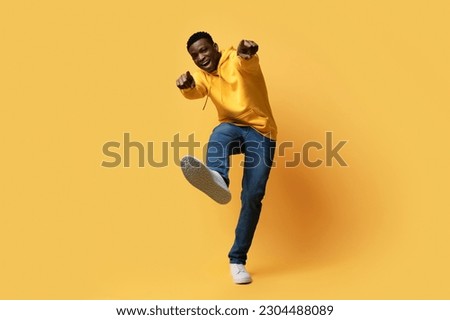 Happy handsome curly young black man in stylish casual outfit showing thumb ups and shoe sneaker sole on yellow studio background, like something or someone, copy space. Human gestures concept