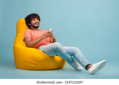 Happy handsome curly young eastern guy in casual outfit chilling at bean bag with smartphone over blue studio background, chatting with friends or playing online games, looking at copy space - Shutterstock ID 2147121047