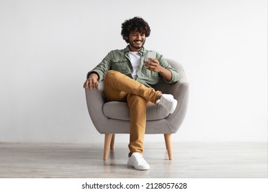 Happy Handsome Curly Young Indian Man Chatting With Girlfriend While Relaxing In Armchair At Home, Using Modern Mobile Phone, Checking Social Media, Using Mobile App, Panorama With Copy Space