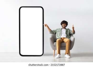 Happy Handsome Curly Young Indian Man Relaxing In Armchair At Home, Pointing At Huge Modern Mobile Phone With White Empty Screen, Recommending Mobile App, Panorama With Copy Space, Mockup