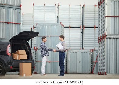 Happy handsome customer in casual clothing shaking hand of storage manager with clipboard after discussing contract about rent of container