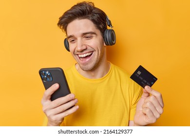 Happy handsome dark haired man types data from credit card in banking app pays easily from home dressed in casual t shirt wears stereo headphones on ears purchases goods in internet poses indoors