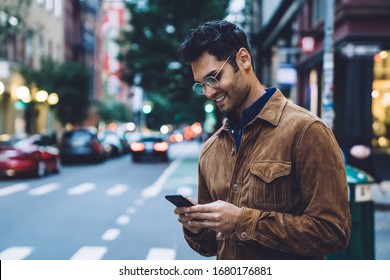 Happy handsome ethnic brunette adult man in jacket and eyeglasses using smartphone while standing beside traffic lane in daylight. Waiting for taxi cab order by application  - Powered by Shutterstock