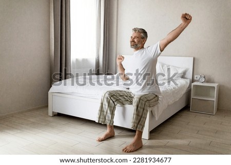 Happy handsome grey-haired bearded middle aged man wearing pajamas sitting on bed and stretching arms, looking at window and smiling, enjoying new sunny day, copy space. Good sleep concept