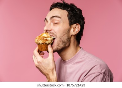 Happy handsome guy eating muffin with eyes closed isolated over pink background
