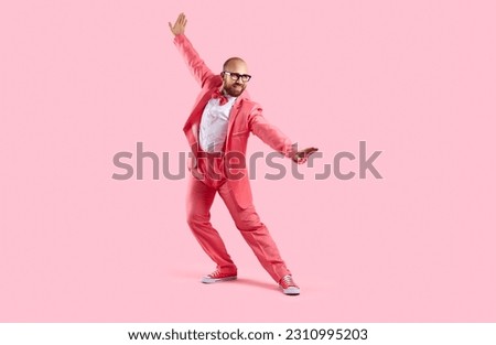 Happy handsome guy in a pink party suit dancing isolated on a pink background. Funny confident bald young man wearing a suit, bowtie, sneakers and glasses dancing and having fun in the studio