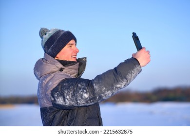 Happy Handsome Guy, Young Man Blogger Is Taking Selfie, Picture Of Himself For Social Media On His Cell Mobile Phone, Looking At Frontal Camera Of Smartphone Outdoors At Winter Cold Frosty Snowy Day