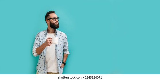 Happy handsome hipster guy dressed in casual clothes in glasses with a beard holds a mug of fragrant coffee or tea in his hand isolated on a blue studio background.
