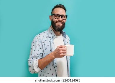 Happy handsome hipster guy dressed in casual clothes in glasses with a beard holds a mug of fragrant coffee or tea in his hand isolated on a blue studio background. - Powered by Shutterstock