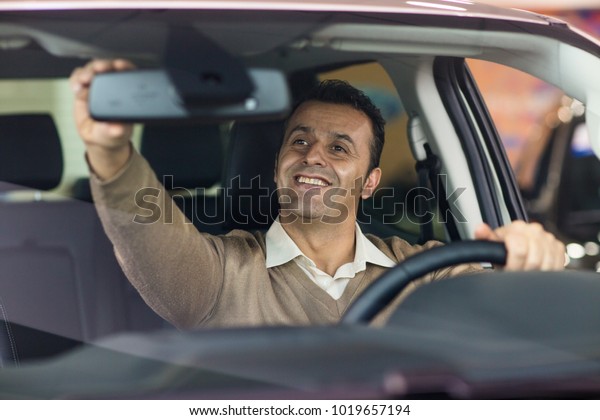 Happy handsome Hispanic mature man smiling\
adjusting rearview mirror sitting in a new car copyspace safety\
driving rental leasing comfort travelling luxury buying vehicle\
driver happiness.