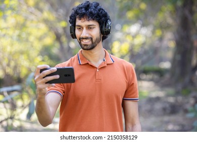 Happy handsome Indian man 30s holding smartphone and wearing headphones in the park outdoor, Young Asian male watching video move or palying game online on his mobile technology concept. phone