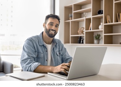 Happy handsome Latin entrepreneur man typing on laptop at office work table, looking at camera with toothy smile, posing for professional portrait, enjoying business success, successful job