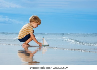 Happy handsome little blond boy put toy boat in the ocean waves at the beach during summer vacation - Shutterstock ID 2233032509